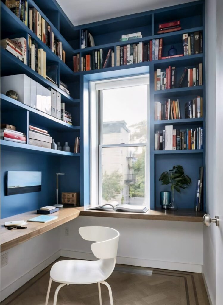 Small home office design with blue wall bookshelves, wooden desk, white chair, and window-side reading nook.